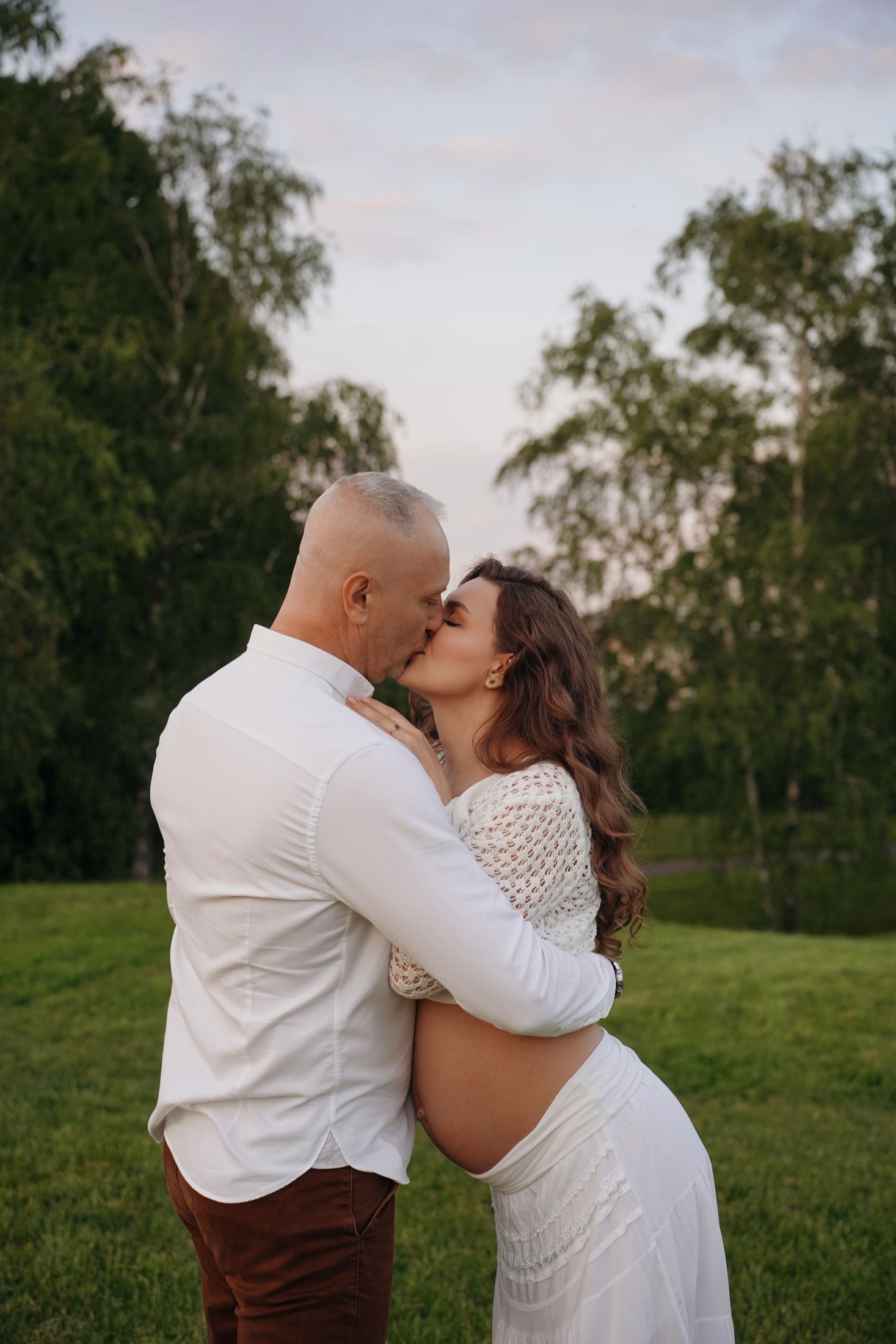 Фотосесія вагітності на природі у Києві на заході сонця, чоловік цілує вагітну жінку у білому одязі