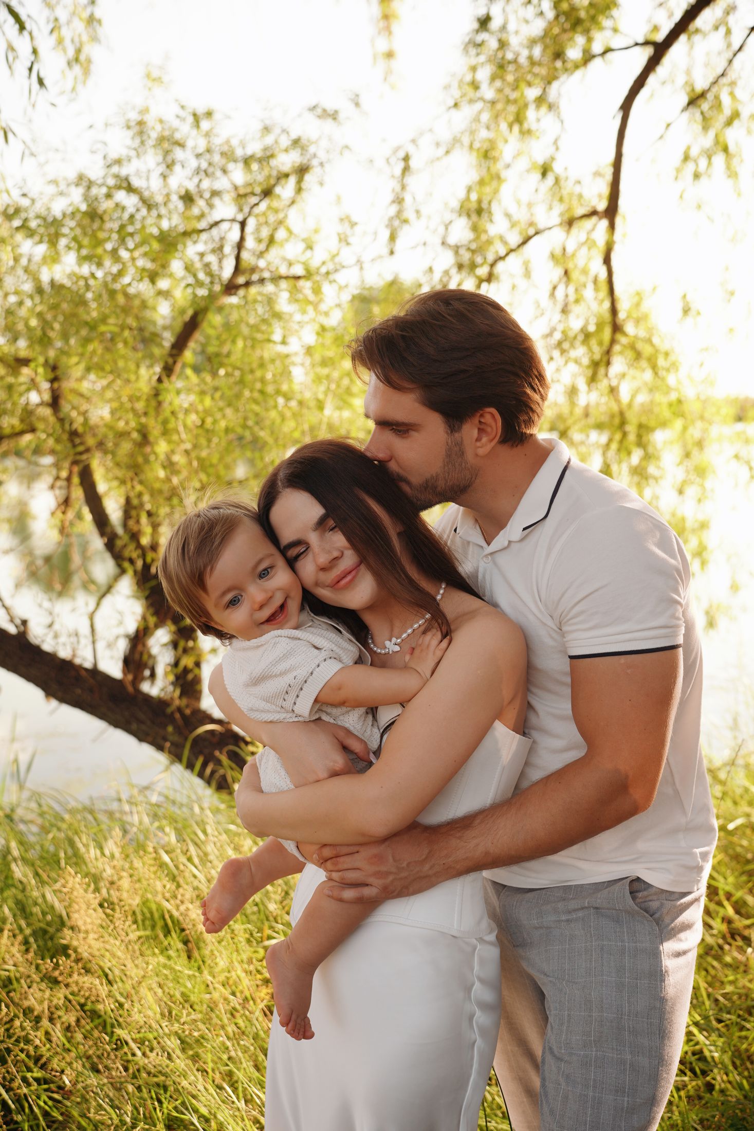 Сімейна фотосесія на природі у Києві - родина обнімається, чоловік цілує жінку, яка тримає на руках усміхнену дитину